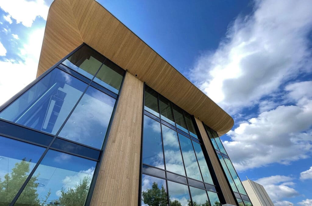 Een modern gebouw met Millboard gevelbekleding en grote glazen ramen die de lucht en wolken reflecteren, onder een blauwe lucht.