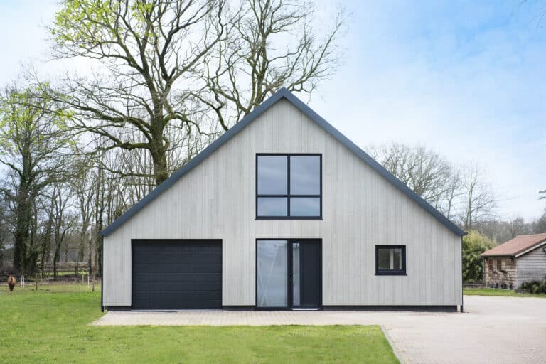 Een moderne schuurwoning met een metalen dak en strakke Millboard gevelbekleding, omgeven door een groene omgeving en klein schuurtje rechts.