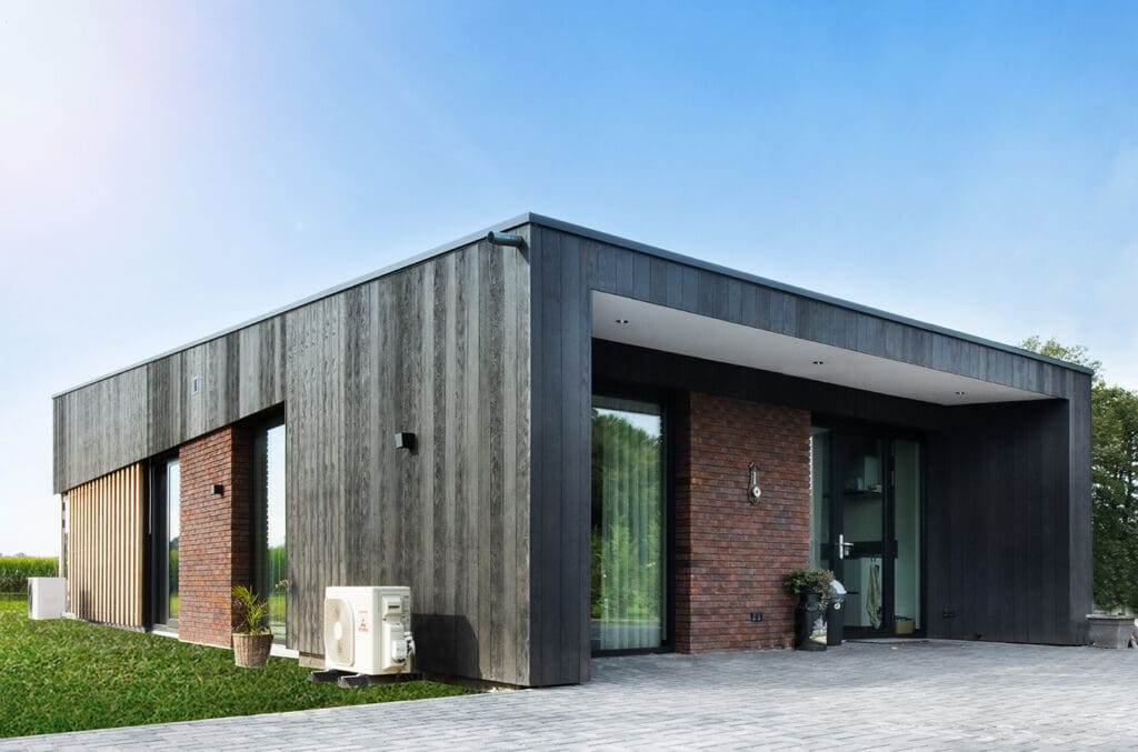 Een modern, laagbouw huis met donkere gevelbekleding en bakstenen accenten, gelegen in een groene omgeving onder de heldere blauwe lucht.