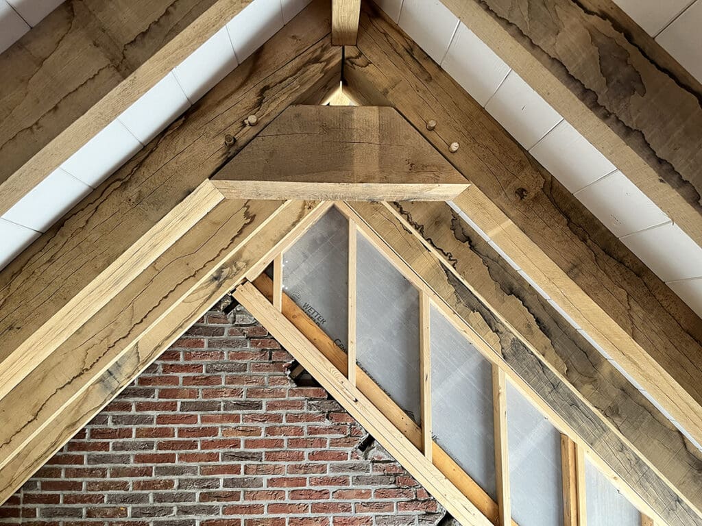 Een binnenaanzicht van een dakconstructie met eiken balken, gevelplaten en een gemetselde bakstenen muur.