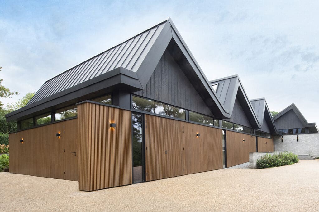Een modern gebouw met zwarte puntdaken en millboard gevelbekleding, voorzien van grote ramen en buitenverlichting, gelegen op een terrein met grind.
